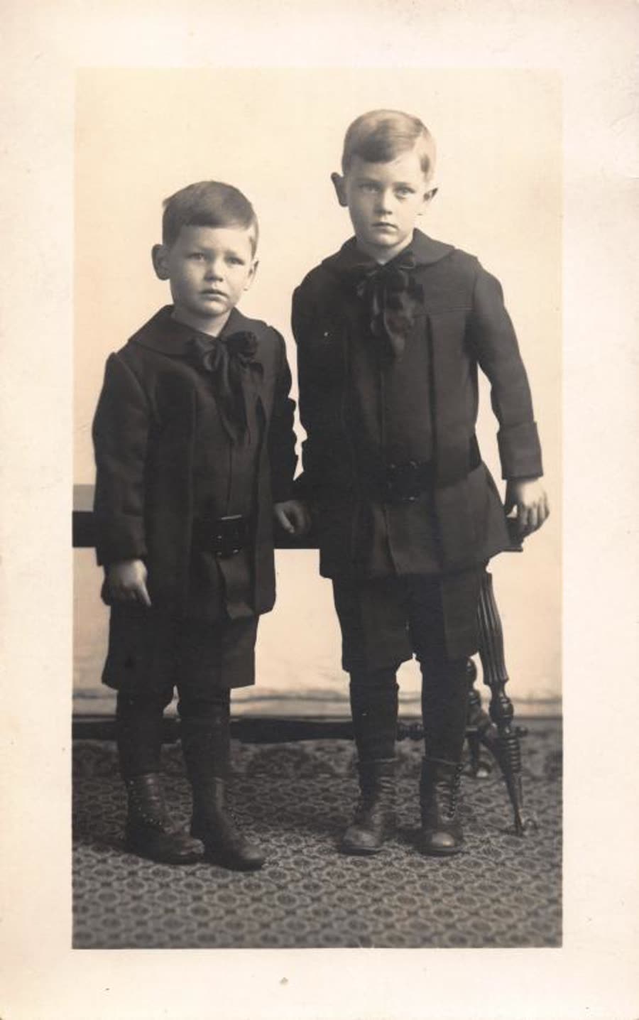 Sepia studio portrait from the 1910s: two young brothers in dark coats with large bows.
