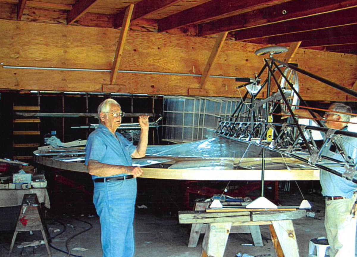 Curtis in blue work clothes hand-building a wood-and-fabric aircraft frame in his plywood workshop.