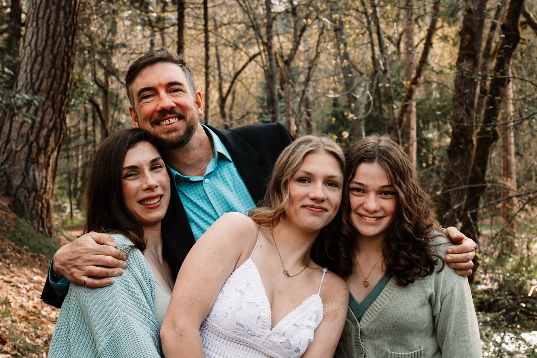Andrew, Amie, Aubrey, and Emily Barnes hugging close together among the pines in spring, everyone smiling.