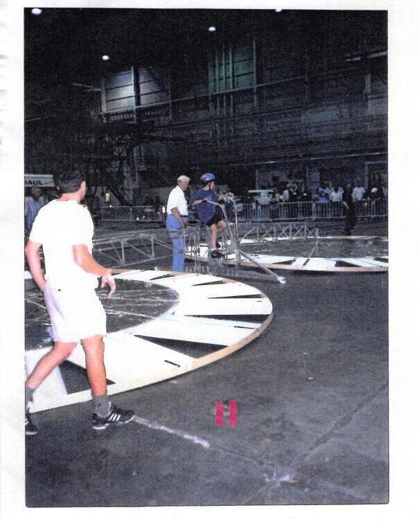 Andrew in a helmet pedals the family machine on the hangar floor, the rotor ring stretching across the frame, while a man strides past in the foreground.