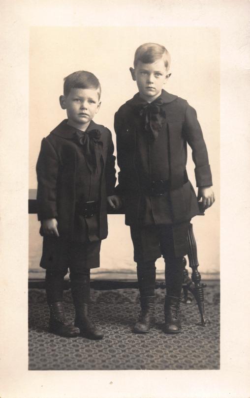 Studio portrait of two young brothers in dark coats and enormous ribbon bows, standing side by side on patterned carpet, unsmiling. About 1925.