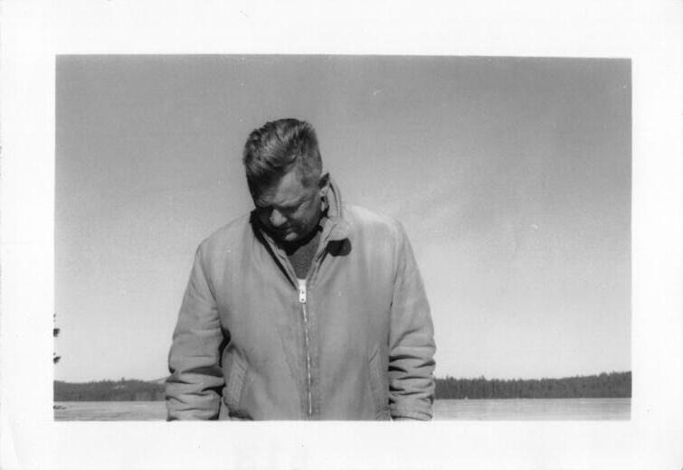 Curtis in a light zip jacket beside a lake in winter, head bowed, his face in shadow. Undated.