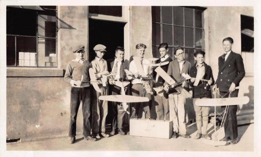 Eight boys and a young man in a suit pose with handmade model gliders outside a workshop window. About 1934.