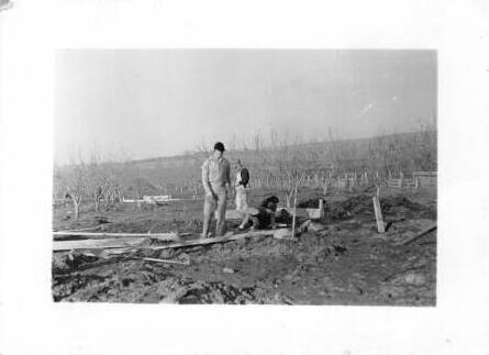 Curtis stands on a plank walkway in a bare, newly worked orchard while another man crouches at an irrigation box. About 1948.