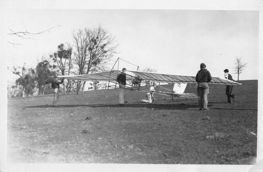 A crew of five carries and steadies a large open-frame glider on a bare hillside, winter trees behind them.