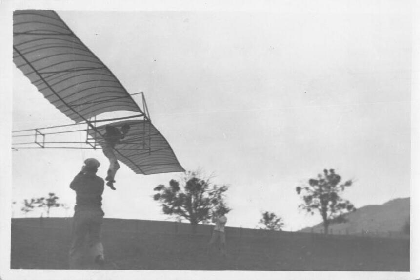 The glider airborne against a white sky, a man hanging beneath its wing, while two people watch from the darkening field below.
