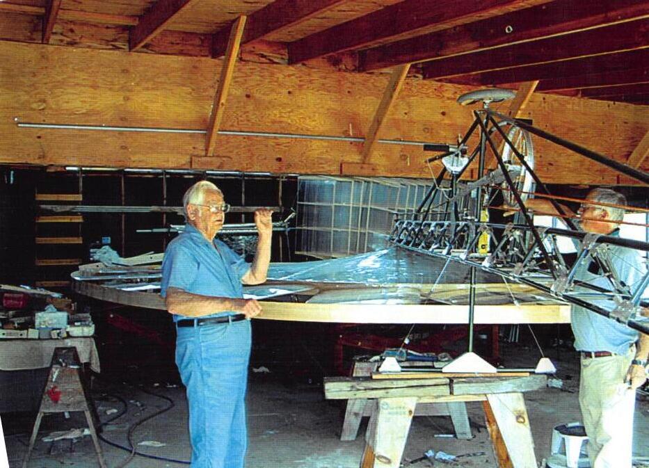 An elderly Curtis in a barn workshop beside the Tipsy Bee: a film-covered rotor nearly the width of the room, with the pedal frame rising at its center. Another man works across the rotor.