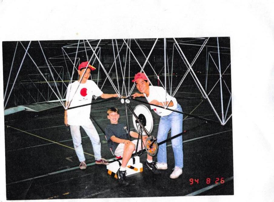 Inside the Boeing hangar, two members of the Japanese team in red caps pose with young Andrew, who sits at the pedals of their truss-framed helicopter. The camcorder date stamp reads 94 8 26.