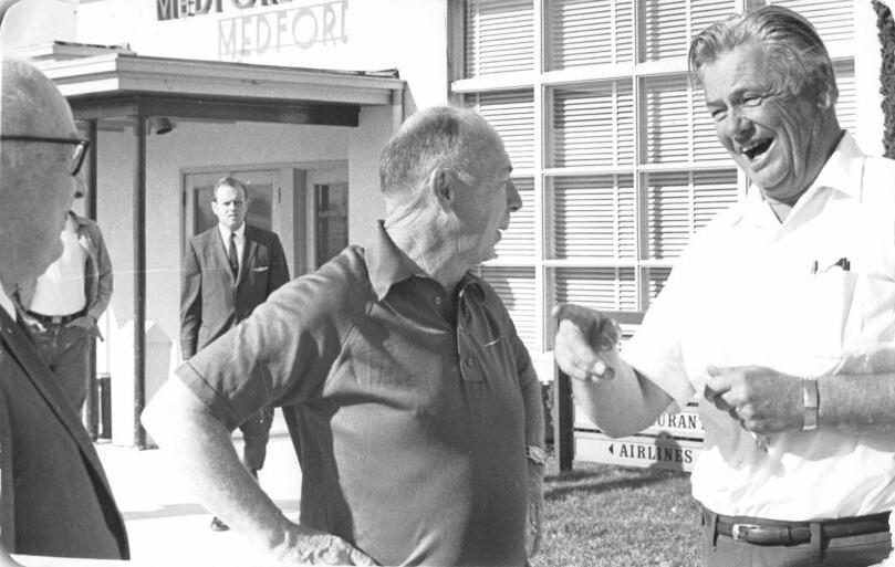 Three men laughing hard outside the Medford airport terminal; an Airlines sign points behind them. About 1965.